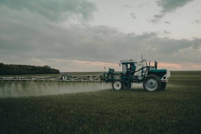 A large agricultural machine is spraying crops in a vast, open field. It is overcast with a cloudy sky, and the landscape is expansive with distant tree lines.