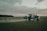 A large agricultural machine is spraying crops in a vast, open field. It is overcast with a cloudy sky, and the landscape is expansive with distant tree lines.