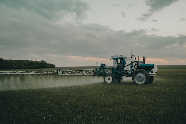 A large agricultural machine is spraying crops in a vast, open field. It is overcast with a cloudy sky, and the landscape is expansive with distant tree lines.