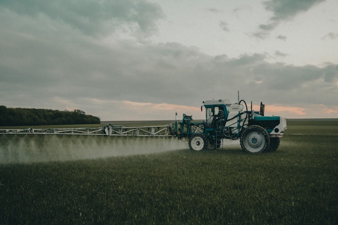 A large agricultural machine is spraying crops in a vast, open field. It is overcast with a cloudy sky, and the landscape is expansive with distant tree lines.
