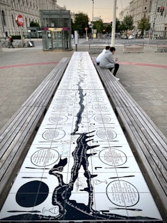 A public space with a large tiled bench featuring artwork and text that runs along its surface. Two people are sitting on one side of the bench, both looking at their phones. The surrounding area includes urban elements such as a tram or metro station sign, bicycles, trees, and a historic building facade.