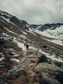 A rugged mountainous landscape with patches of snow and a rocky trail. A group of people is hiking up the trail surrounded by steep slopes. The sky is overcast, lending a moody atmosphere to the scene.