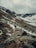 A rugged mountainous landscape with patches of snow and a rocky trail. A group of people is hiking up the trail surrounded by steep slopes. The sky is overcast, lending a moody atmosphere to the scene.