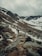 A rugged mountainous landscape with patches of snow and a rocky trail. A group of people is hiking up the trail surrounded by steep slopes. The sky is overcast, lending a moody atmosphere to the scene.