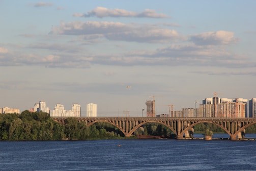 A river flowing under a large, arched bridge with multiple lanes. High-rise buildings and construction cranes are visible in the background, suggesting urban development. Lush green trees line the riverbank, and the sky is mostly clear with scattered clouds.