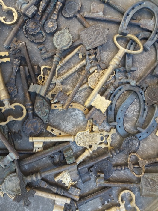 A variety of ornate, vintage keys and horseshoes scattered across a flat surface. The keys are intricately designed, featuring detailed engravings and different shapes. The collection includes metals with a patina, indicating age, with some bronze and dark steel colors.