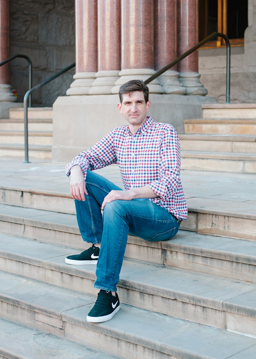 man in blue and white checkered dress shirt and blue denim jeans sitting on brown concrete
