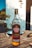 A bottle of Havana Club Añejo Especial Cuban rum is placed on a wooden outdoor table. The setting suggests a sunny day, with blurry background elements indicating an outdoor dining area. The label on the bottle features distinctive branding and blue and gold accents. Other objects, such as a utensil and a plate, are partially visible, emphasizing a casual and relaxed outdoor atmosphere.