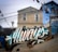 A house is in the background with some visible windows, surrounded by various colorful graffiti and murals on the nearby walls. The word 'always' is prominently painted in white across a dark-colored wall in the foreground, accented with teal and gold colors. There are many power lines running overhead and a streetlamp. The ground is paved with cobblestones, and there are two small, wooden dog houses along with a dog lying near them.