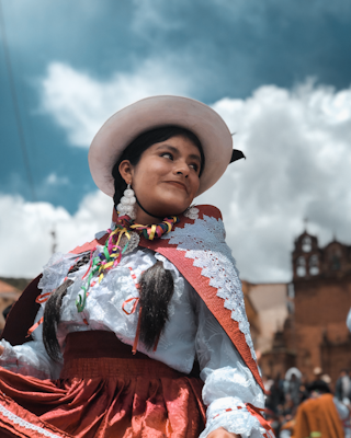 A person is wearing traditional attire with a white hat and colorful earrings. The garment includes a red skirt and a lace-trimmed cape. The background features an outdoor setting with a partly cloudy sky and historical architecture.