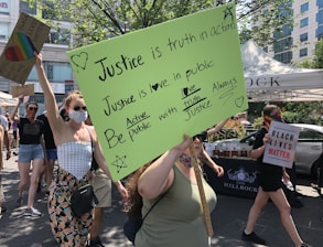 A group of people is participating in a protest or rally. They are holding various signs, one of which has a bright green background with handwritten messages about justice and love. Another sign reads 'Black Lives Matter.' One person is wearing a rainbow mask, adding to the message of inclusivity and diversity. The scene is set outdoors under bright daylight, with trees and a building visible in the background.