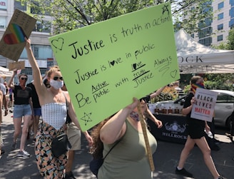 A group of people is participating in a protest or rally. They are holding various signs, one of which has a bright green background with handwritten messages about justice and love. Another sign reads 'Black Lives Matter.' One person is wearing a rainbow mask, adding to the message of inclusivity and diversity. The scene is set outdoors under bright daylight, with trees and a building visible in the background.