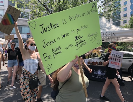 A group of people is participating in a protest or rally. They are holding various signs, one of which has a bright green background with handwritten messages about justice and love. Another sign reads 'Black Lives Matter.' One person is wearing a rainbow mask, adding to the message of inclusivity and diversity. The scene is set outdoors under bright daylight, with trees and a building visible in the background.