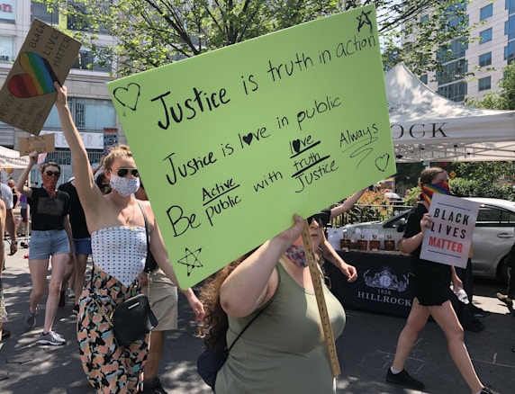 A group of people is participating in a protest or rally. They are holding various signs, one of which has a bright green background with handwritten messages about justice and love. Another sign reads 'Black Lives Matter.' One person is wearing a rainbow mask, adding to the message of inclusivity and diversity. The scene is set outdoors under bright daylight, with trees and a building visible in the background.