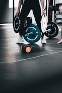 A person lifting a heavy barbell in a gym setting, using both hands to stabilize the weights attached to either end. The floor is marked with lines, and a water bottle lies nearby. The focus is on the person's lower body and the weights.