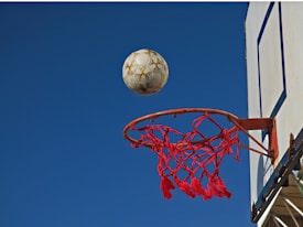 A basketball net is positioned under a clear blue sky. A soccer ball is in mid-air above the hoop, appearing to be on the verge of falling through. The netting of the hoop is red, while the backboard is white with blue detailing.