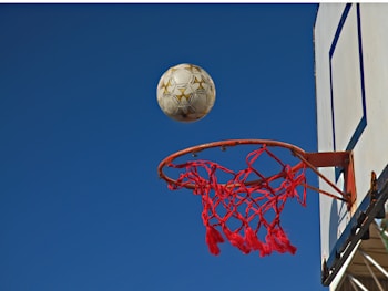 A basketball net is positioned under a clear blue sky. A soccer ball is in mid-air above the hoop, appearing to be on the verge of falling through. The netting of the hoop is red, while the backboard is white with blue detailing.