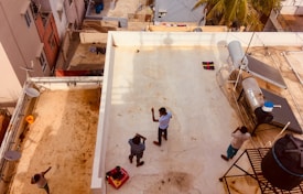 A view from above shows a group of people on a rooftop, one person appears to be engaged in a physical activity or exercise while others watch or take photos. The rooftop is equipped with a solar water heater and other utility structures, like satellite dishes. The surrounding buildings are visible, along with electrical wires and a palm tree in the distance.