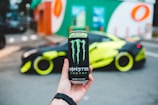 A hand holding a can of Monster Energy drink, prominently displayed in the foreground, with a bright, customized sports car parked in the background. The car has neon yellow accents and is parked near a building with colorful signage.