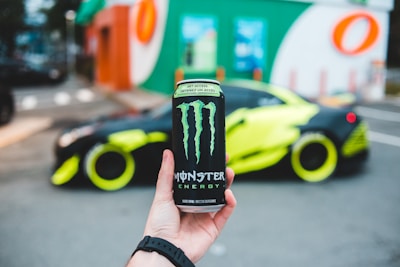 A hand holding a can of Monster Energy drink, prominently displayed in the foreground, with a bright, customized sports car parked in the background. The car has neon yellow accents and is parked near a building with colorful signage.
