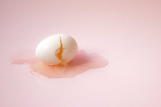A cracked egg with a yolk visible is resting on a flat pink surface, surrounded by spilled egg white.