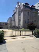 A large, multi-story stone building with distinct architectural features such as bay windows and a steeply pitched roof with dormer windows. The building is situated behind a black metal fence, which borders a driveway or entryway with neatly trimmed bushes and greenery. The scene is set on a clear day with a bright blue sky in the background.