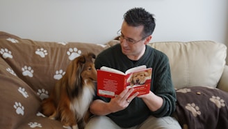 man in sweater holding a book, looking at brown and white dog