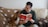 A man with short, dark hair wearing glasses and a green sweater sits on a beige couch with a brown blanket featuring white paw prints. He is reading a red book to a sable and white Shetland Sheepdog sitting beside him, which is attentively looking at the book.