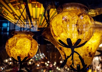Two intricately designed stained glass lamps with warm amber hues and dark metallic accents in the shape of dragonflies. In the background, a larger stained glass fixture with geometric patterns and earth tones is partially visible, creating a warm, inviting ambiance with reflections and subtle lighting effects.