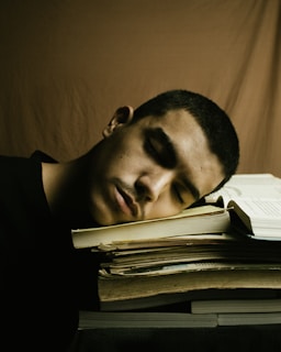 A young man appears to be asleep with his head resting on a stack of books. The background is a plain brown fabric, and a book is open in front of him.