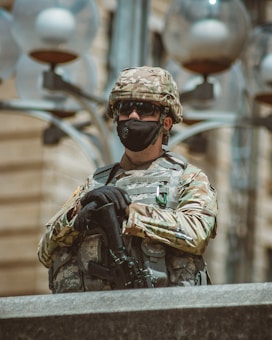A soldier wearing camouflage uniform and tactical gear stands guard, holding a firearm. The individual is equipped with a helmet and black face mask. The background shows part of a building and several round, opaque lamp structures.