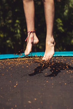 A pair of feet are suspended above a trampoline, with leaves and debris scattered around, suggesting a playful jumping motion. A colorful anklet is visible on one foot, adding a festive touch to the scene. The background consists of blurred greenery.