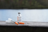 A vial of insulin and two syringes are placed on a wooden surface. The background features a blurred view of a lake and greenery, giving the scene a peaceful and serene outdoor setting.