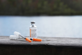 A vial of insulin and two syringes are placed on a wooden surface. The background features a blurred view of a lake and greenery, giving the scene a peaceful and serene outdoor setting.