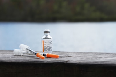 A vial of insulin and two syringes are placed on a wooden surface. The background features a blurred view of a lake and greenery, giving the scene a peaceful and serene outdoor setting.