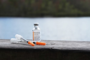 A vial of insulin and two syringes are placed on a wooden surface. The background features a blurred view of a lake and greenery, giving the scene a peaceful and serene outdoor setting.