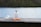 A vial of insulin and two syringes are placed on a wooden surface. The background features a blurred view of a lake and greenery, giving the scene a peaceful and serene outdoor setting.