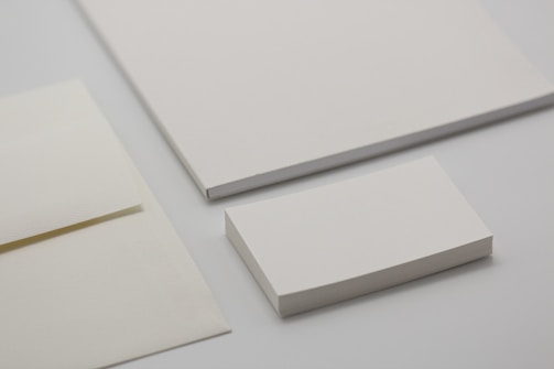 Various forms of blank stationery items are arranged, including a stack of business cards, an envelope, and sheets of paper, all placed on a neutral-colored surface.