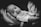 A black and white portrait of an infant being gently cradled in an adult's hand, showcasing a calm and serene expression. The image highlights the soft facial features of the baby and the tenderness of the moment.