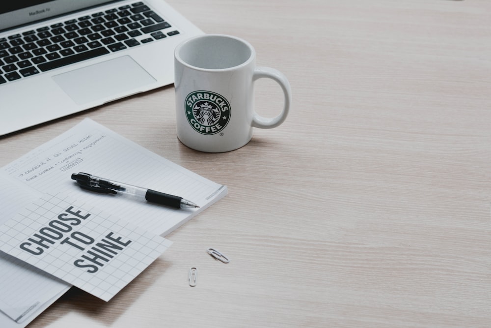 white and black ceramic mug beside black pen on white printer paper