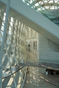 An interior view featuring a modern architectural space with intricate lattice patterns on the side. The design includes a curved pathway and large glass windows allowing natural light to filter through. The space is spacious, with a few people seated in the background.