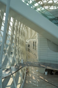 An interior view featuring a modern architectural space with intricate lattice patterns on the side. The design includes a curved pathway and large glass windows allowing natural light to filter through. The space is spacious, with a few people seated in the background.