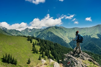 A close-up of a backpacker adjusting their gear on a mountain ridge with a panoramic view of peaks under a clear sky.