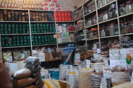 A bustling shop interior filled with packed shelves, showcasing a variety of products such as jars, cans, and bags filled with different grains and spices. Labels with handwritten prices are visible, adding a local market atmosphere. The display is well-organized, with colorful packaging and a diverse selection of goods.