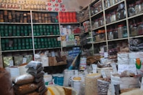 A bustling shop interior filled with packed shelves, showcasing a variety of products such as jars, cans, and bags filled with different grains and spices. Labels with handwritten prices are visible, adding a local market atmosphere. The display is well-organized, with colorful packaging and a diverse selection of goods.