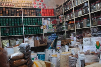 A bustling shop interior filled with packed shelves, showcasing a variety of products such as jars, cans, and bags filled with different grains and spices. Labels with handwritten prices are visible, adding a local market atmosphere. The display is well-organized, with colorful packaging and a diverse selection of goods.