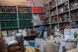 A bustling shop interior filled with packed shelves, showcasing a variety of products such as jars, cans, and bags filled with different grains and spices. Labels with handwritten prices are visible, adding a local market atmosphere. The display is well-organized, with colorful packaging and a diverse selection of goods.