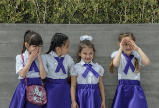 Four young girls in matching outfits stand in front of a wooden fence. Each girl wears a white blouse with lace details and a purple skirt. They all have purple ribbons tied at their collars. One girl is holding a Hello Kitty bag, and they seem to be interacting playfully with each other. Green foliage is visible in the background.