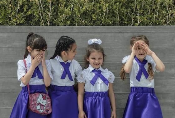 Four young girls in matching outfits stand in front of a wooden fence. Each girl wears a white blouse with lace details and a purple skirt. They all have purple ribbons tied at their collars. One girl is holding a Hello Kitty bag, and they seem to be interacting playfully with each other. Green foliage is visible in the background.