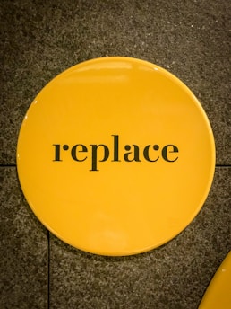 A circular yellow sign with the word 'replace' written in black lowercase letters. The sign is placed on a textured gray surface.